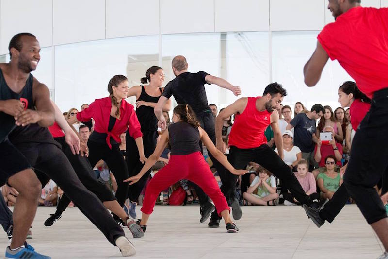 Dancers dressed in black and red smile while dancing in a large group